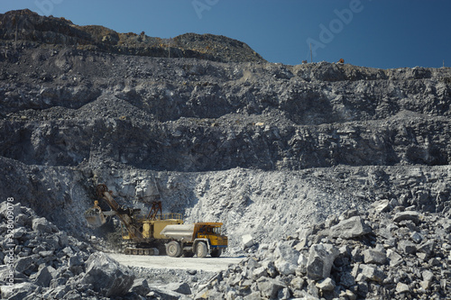 Mining excavator and heavy mining truck in a quarry for the extraction of limestone on the background of rocky terrain and blue sky in sunny weather.