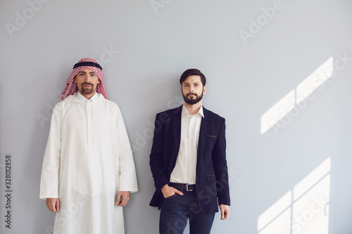 Arab and european businessmen stand smiling at the gray background.