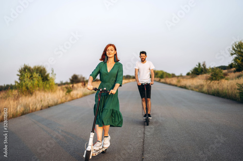 Young happy smiling couple having fun driving electric scooter on the road in the countryside. Content technologies