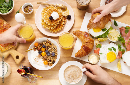 Top view image of brunch menu on wooden table with hands holding food and drinks. Healthy sunday breakfast with croissants, waffles, granola and sandwiches.