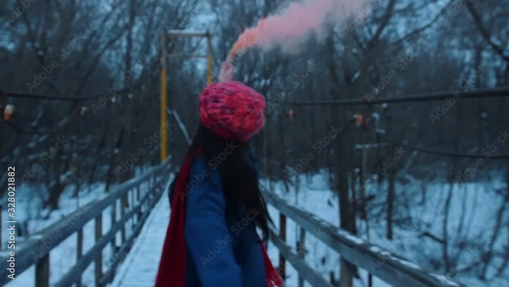 A young smiling woman running on the snowy bridge holding a peachy smoke bomb A young smiling woman running on the snowy bridge holding a peachy smoke bomb