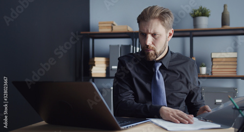 Fotografie Adult concentrated man working with document and looking at laptop screen sittin