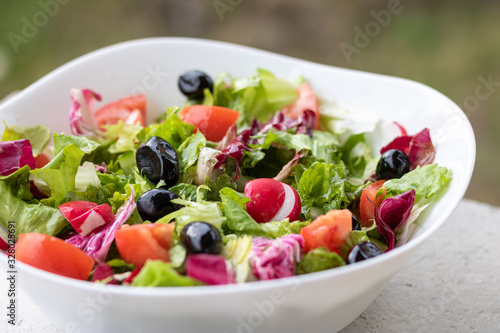 Green vegan salad from green leaves mix and vegetables in white bowl.Summer Easter diet salad dish of lettuce, radicchio, Lollo Rossa, endives, radishes, green onions, cucumbers, olives and tomatoes