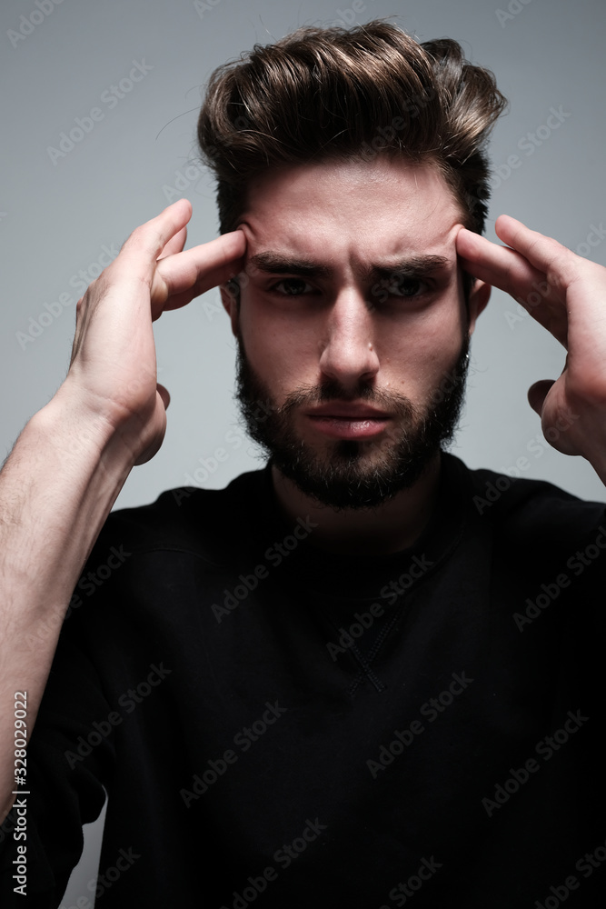 A young dark-haired man with a beard holds his head and shows different human emotions: hatred, fear, despair, horror, malaise, headache, clairvoyance. Close-up studio portrait of a man. A young dark-haired man with a beard holds his head and shows different human emotions: hatred, fear, despair, horror, malaise, headache, clairvoyance. Close-up studio portrait of a man.