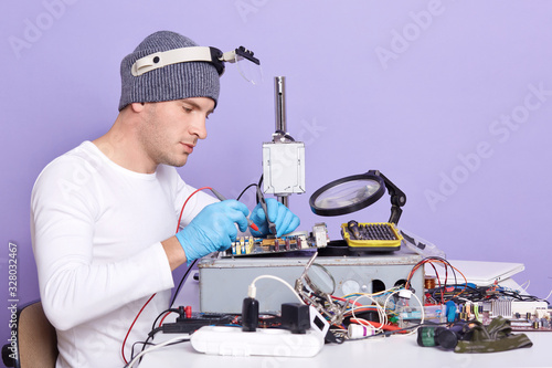 Indoor shot of repairman using magnifier and multimeter while fixing pc motherboard, profile of radioman wears white shirt, gray cap and blue rabber gloves, posing isolated on lilac studio background. photo