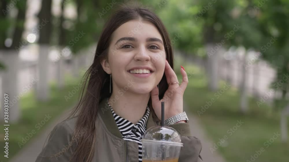 Close up of beautiful brown haired young woman smiling drinking juice beverage, smoothing hair, enjoying warm day relaxed urban lifestyle city street background dressed as stylish millennial hipster Close up of beautiful brown haired young woman smiling drinking juice beverage, smoothing hair, enjoying warm day relaxed urban lifestyle city street background dressed as stylish millennial hipster