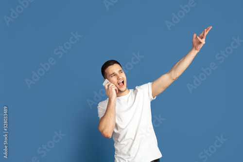 Talking on phone, pointing. Caucasian young man's portrait on blue studio background. Beautiful male model in casual style, pastel colors. Concept of human emotions, facial expression, sales, ad.