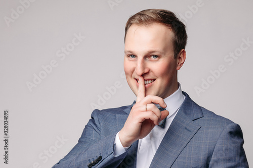 Front view of funny man wearing white shirt and bow tie looking aside and showing sign of pistol on blue isolated background. Young male gesturing and posing in studio. Concept of foolishness.