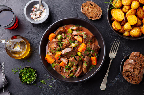 Beef meat and vegetables stew in black bowl with roasted baby potatoes. Dark background. Top view. photo