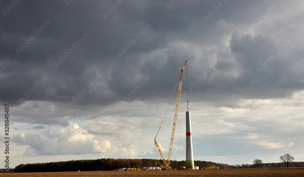 Fototapeta premium Construction of a wind turbine with an enormous crane, dramatic grey clouds in the background