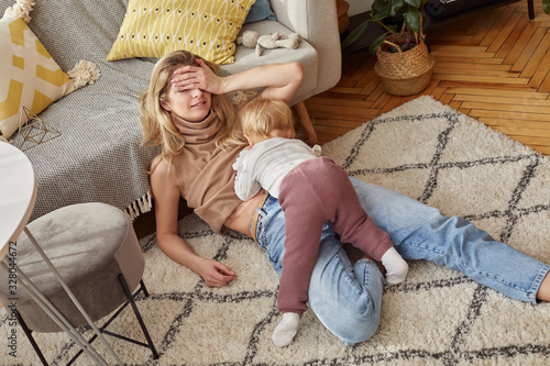 Blonde Caucasian woman in jeans is sitting on carpet her little baby child in a bright room with windows in Scandinavian style breast feeding exhausted