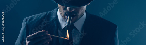 Mafioso lighting cigar with match on dark blue, panoramic shot photo