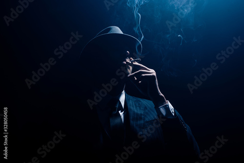 Silhouette of mafioso in suit and felt hat smoking cigarette on dark blue background