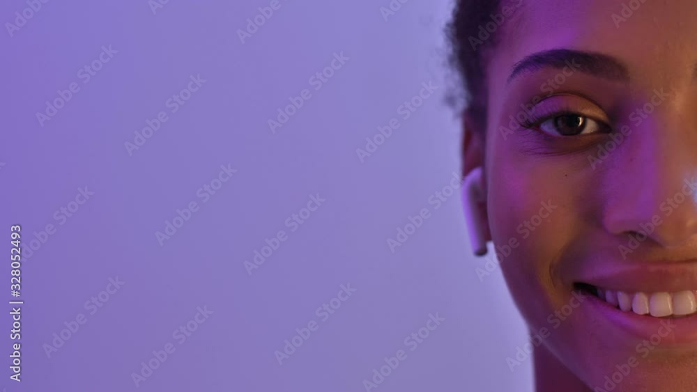 Half face of Smiling african woman in earphones listening music and looking to the camera isolated over purple neon background Half face of Smiling african woman in earphones listening music and looking to the camera isolated over purple neon background