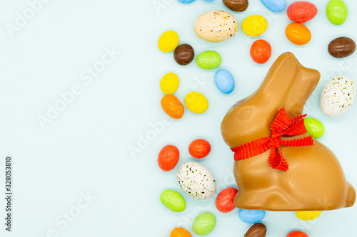 Happy easter card. Easter milk chocolate bunny, rabbit with red bow on a neck. There are colourful candies and small eggs around. Blue background. Isolated. Copy space on the left side.
