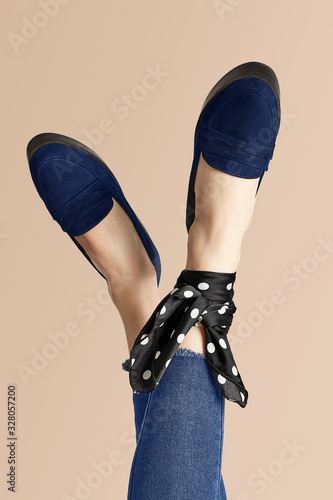 Close-up shot of crossed female legs in blue loafers and jeans on the beige background. One of the anklesis wrapped with a black silk handkerchief with white polka-dot print.  photo