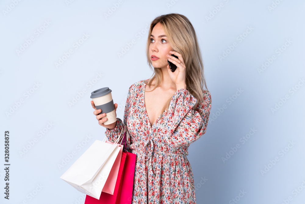 Teenager Russian girl with shopping bag isolated on blue background holding coffee to take away and a mobile Teenager Russian girl with shopping bag isolated on blue background holding coffee to take away and a mobile