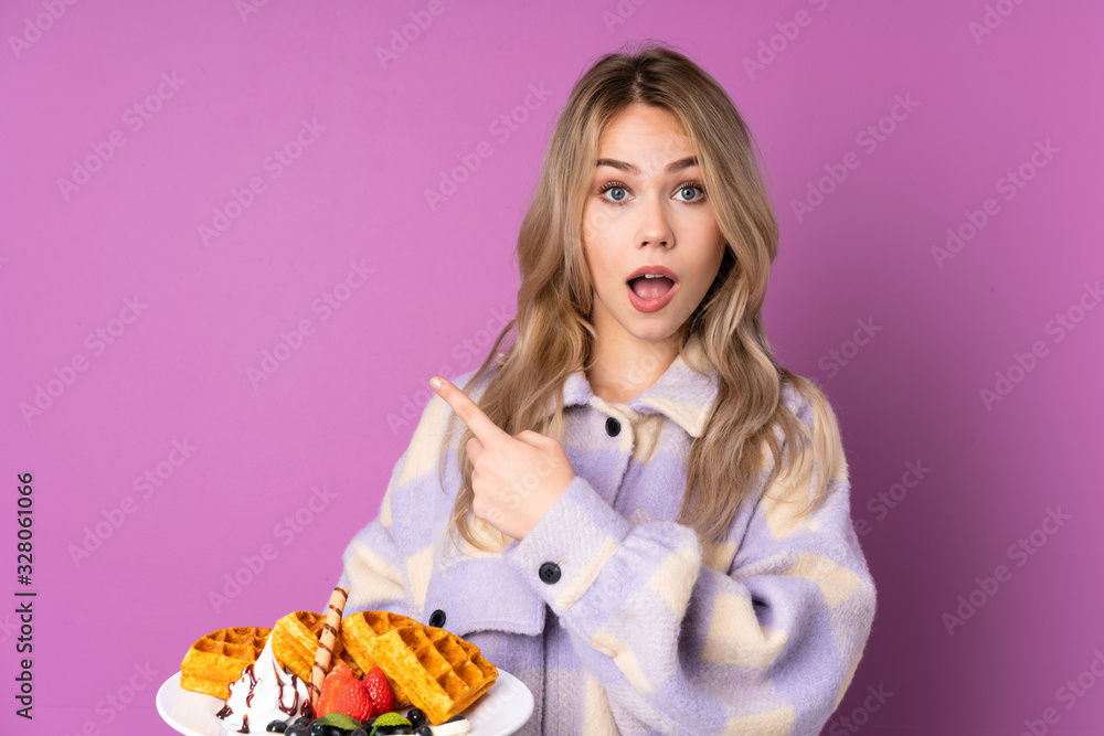 Teenager Russian girl holding waffles isolated on purple background surprised and pointing side Teenager Russian girl holding waffles isolated on purple background surprised and pointing side