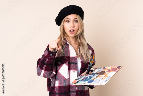 Teenager artist girl holding a palette isolated on blue background surprised and pointing front