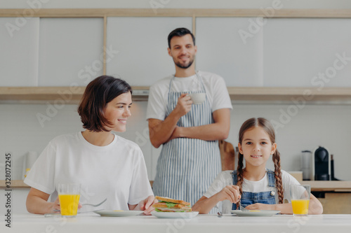 Lovely small kid and mother have breakfast together  sit at kitchen table  eat delicious meal  father stands in background  wears apron and drink coffee. Friendly family gather together at kitchen