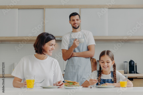 Happy mother talks to daughter while have breakfast. Father stands behind  prepared delicious dish for family. Friendly family members meet at kitchen during weekend  enjoy nice conversation