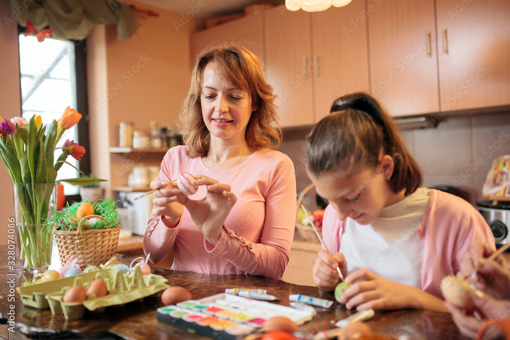 Fototapeta premium Smiling happy mid adult Caucasian woman decorating and painting Easter eggs and having a great time with her teenage daughter at home