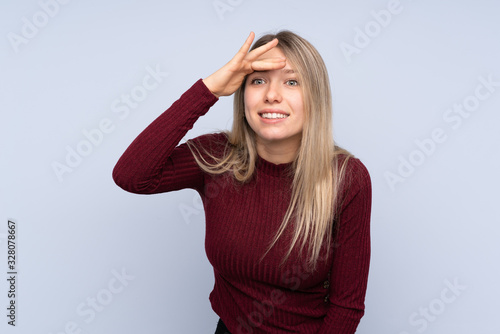 Young blonde woman over isolated blue background looking far away with hand to look something