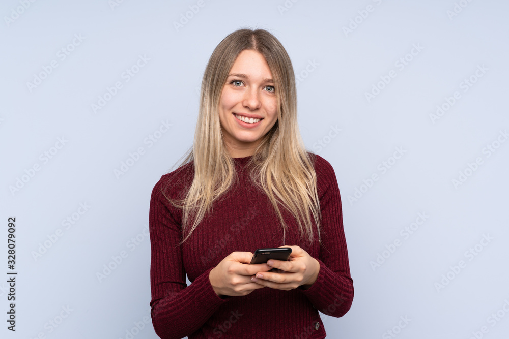 Young blonde woman over isolated blue background sending a message with the mobile Young blonde woman over isolated blue background sending a message with the mobile
