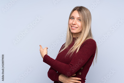 Young blonde woman over isolated blue background pointing back