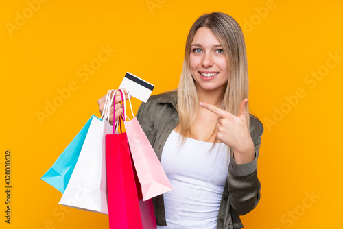 Young blonde woman over isolated yellow background holding shopping bags and a credit card