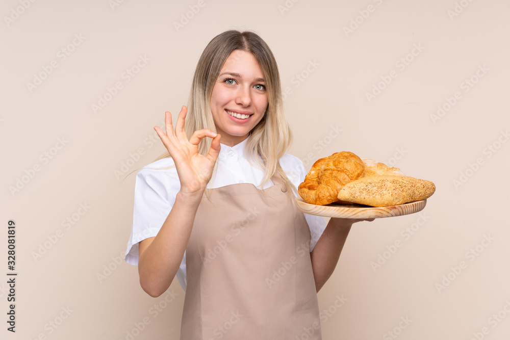 Young blonde girl with apron. Female baker holding a table with several breads showing ok sign with fingers Young blonde girl with apron. Female baker holding a table with several breads showing ok sign with fingers