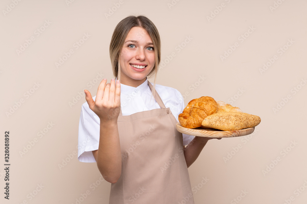 Young blonde girl with apron. Female baker holding a table with several breads inviting to come with hand. Happy that you came