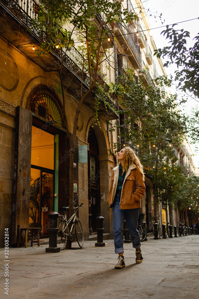 Blond girl walking through the streets of the center of the city of Barcelona in Catalonia, Spain wearing a jacket and blue jeans. Looking up to the old buldings. Blond girl walking through the streets of the center of the city of Barcelona in Catalonia, Spain wearing a jacket and blue jeans. Looking up to the old buldings.
