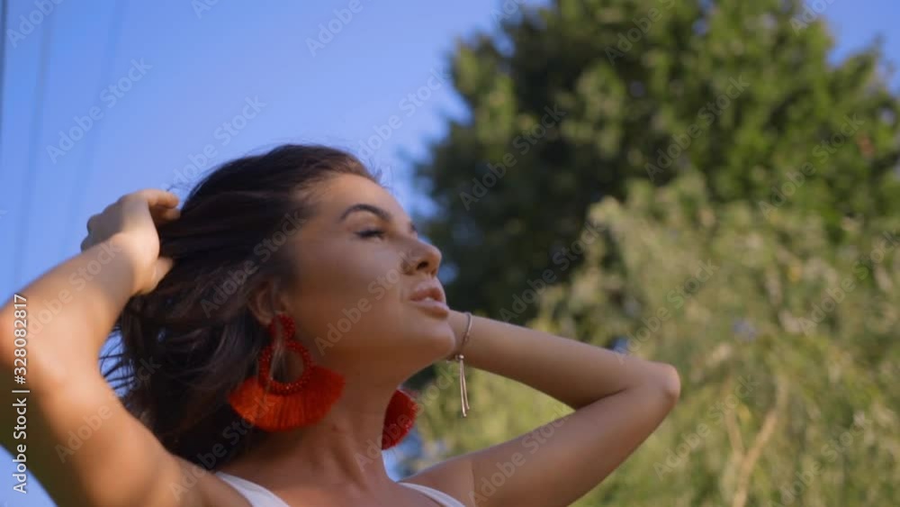 Beautiful girl touches her shiny curly hair and slowly walks in bright rays. Attractive young woman enjoys summer sun and breathes the fresh air. Sexy tanned brunette model in red earrings outdoors. Beautiful girl touches her shiny curly hair and slowly walks in bright rays. Attractive young woman enjoys summer sun and breathes the fresh air. Sexy tanned brunette model in red earrings outdoors.
