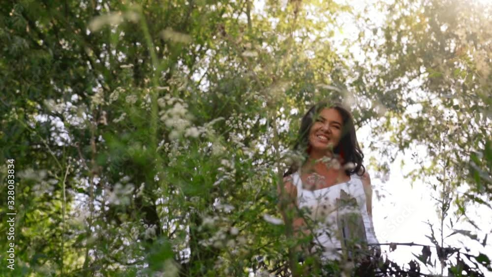 Tanned young woman with sexy lips and curly hair enjoys summer nature and sunlight in a village. Beautiful brunette smiles, stretches and slowly moves in green grass among trees. Model poses outdoors.