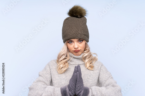 Young teenager girl with winter hat over isolated blue background pleading © luismolinero