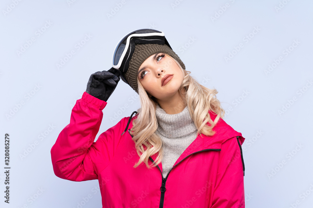 Skier teenager girl with snowboarding glasses over isolated blue background having doubts and with confuse face expression