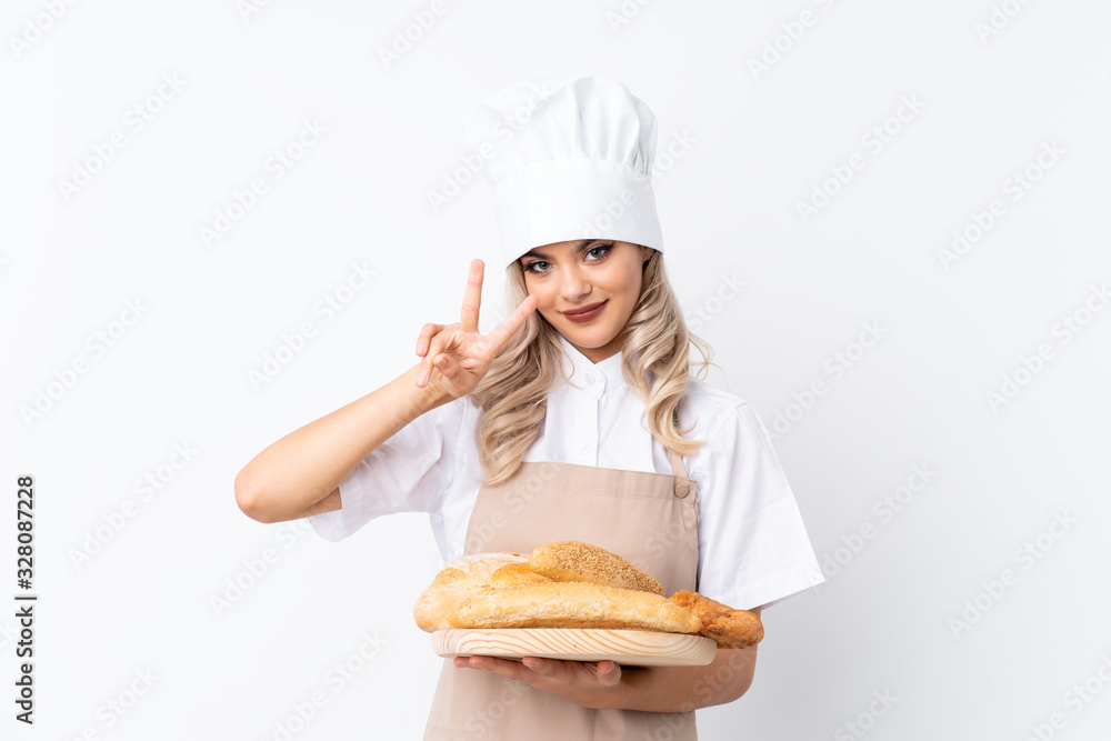 Teenager girl in chef uniform. Female baker holding a table with several breads over isolated white background smiling and showing victory sign