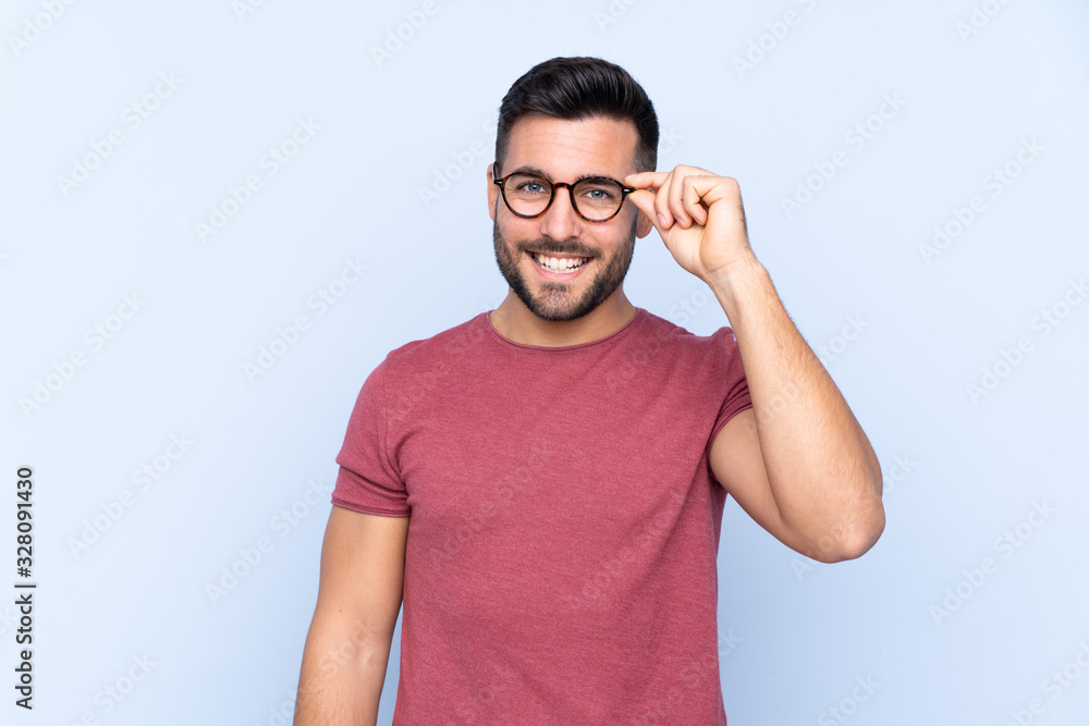 Young handsome man with beard over isolated blue background with glasses and happy Young handsome man with beard over isolated blue background with glasses and happy