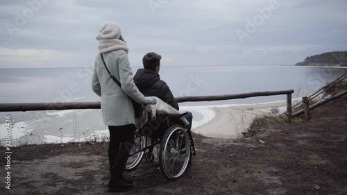 Back side view of woman standing with wheelchair with disabled boy in it. Caucasian crippled man and mother or invalid tender spending autumn day on river bank outdoors. Support, care, disability.