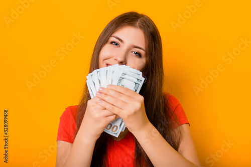Portrait of a smiling young woman holding a bunch of money banknotes in hands isolated on a yellow background.