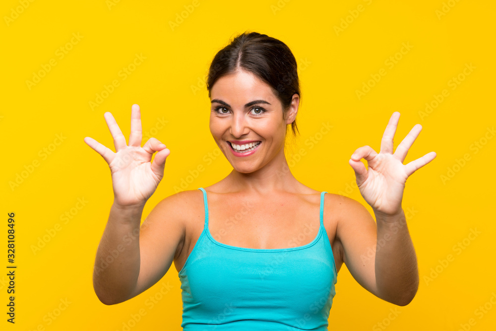 Young woman over isolated yellow background showing an ok sign with fingers Young woman over isolated yellow background showing an ok sign with fingers