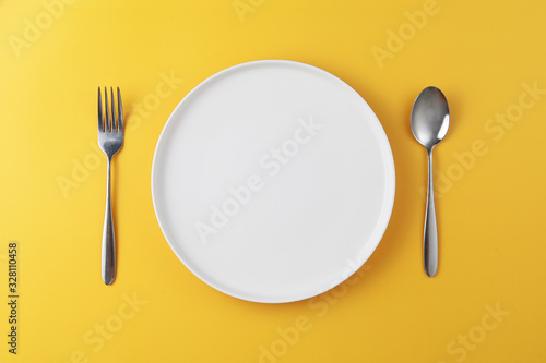 Empty white plate with spoon and fork on yellow background