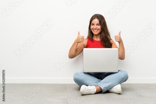 Young woman with a laptop sitting on the floor giving a thumbs up gesture