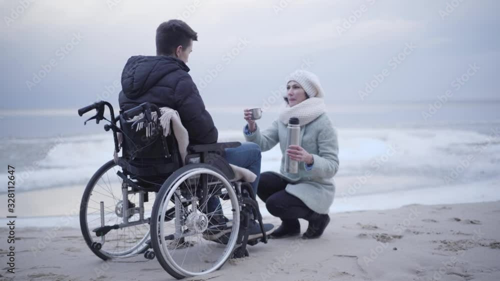 Beautiful Caucasian woman pouring tea from thermos bottle and giving it to disabled boy in wheelchair. Smiling invalid tender or mother taking care of crippled young man on autumn sea shore. Beautiful Caucasian woman pouring tea from thermos bottle and giving it to disabled boy in wheelchair. Smiling invalid tender or mother taking care of crippled young man on autumn sea shore.