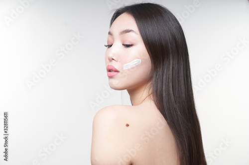 Portrait of a young Asian girl with a horizontal stripe on her cheek, smeared with white cream. Looks down at the floor. On white background. The concept of skin care and beauty 
