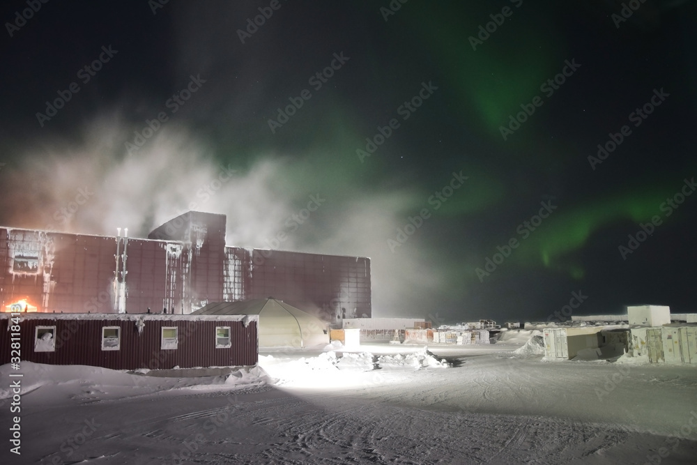 Fototapeta premium Blizzard snow storm and Northern Lights dancing over an ore processing plant in arctic region (Canada)