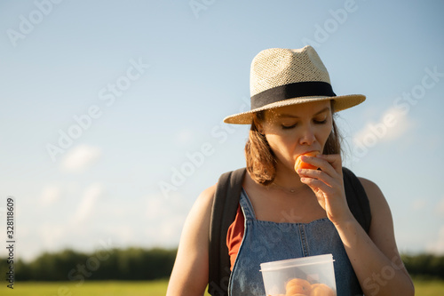 Young woman in hat standing in field eating apricot 