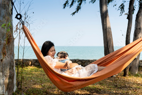 Funny cute pug dog smiling and tongue sticking out while on a trip with her owner, joyful young family, woman lying rest in comfortable hammock on sea beach. Happy times concept.