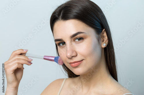 Close-up of a young girl in a light top on a white background making a facial make-up. A pretty woman holds a cosmetic brush near her face and smiles.